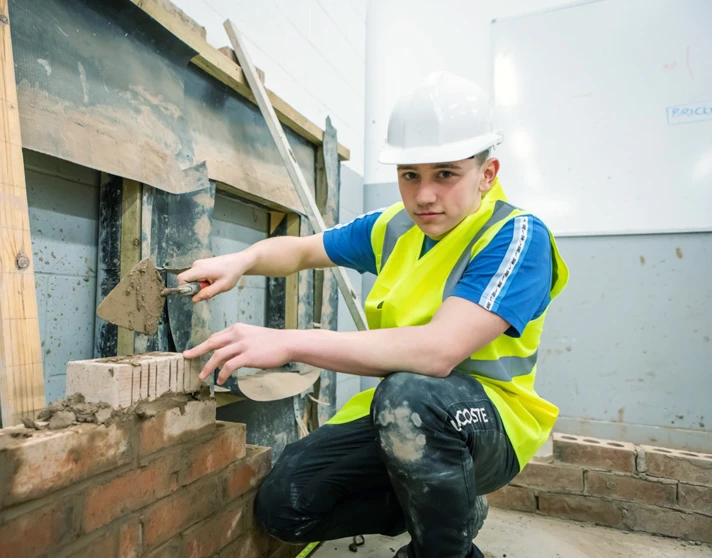 A brickwork student wearing a safety helmet and high-visibility vest crouching down to carefully apply mortar to bricks using a trowel during practical training. A brickwork student wearing a safety helmet and high-visibility vest crouching down to carefully apply mortar to bricks using a trowel during practical training.